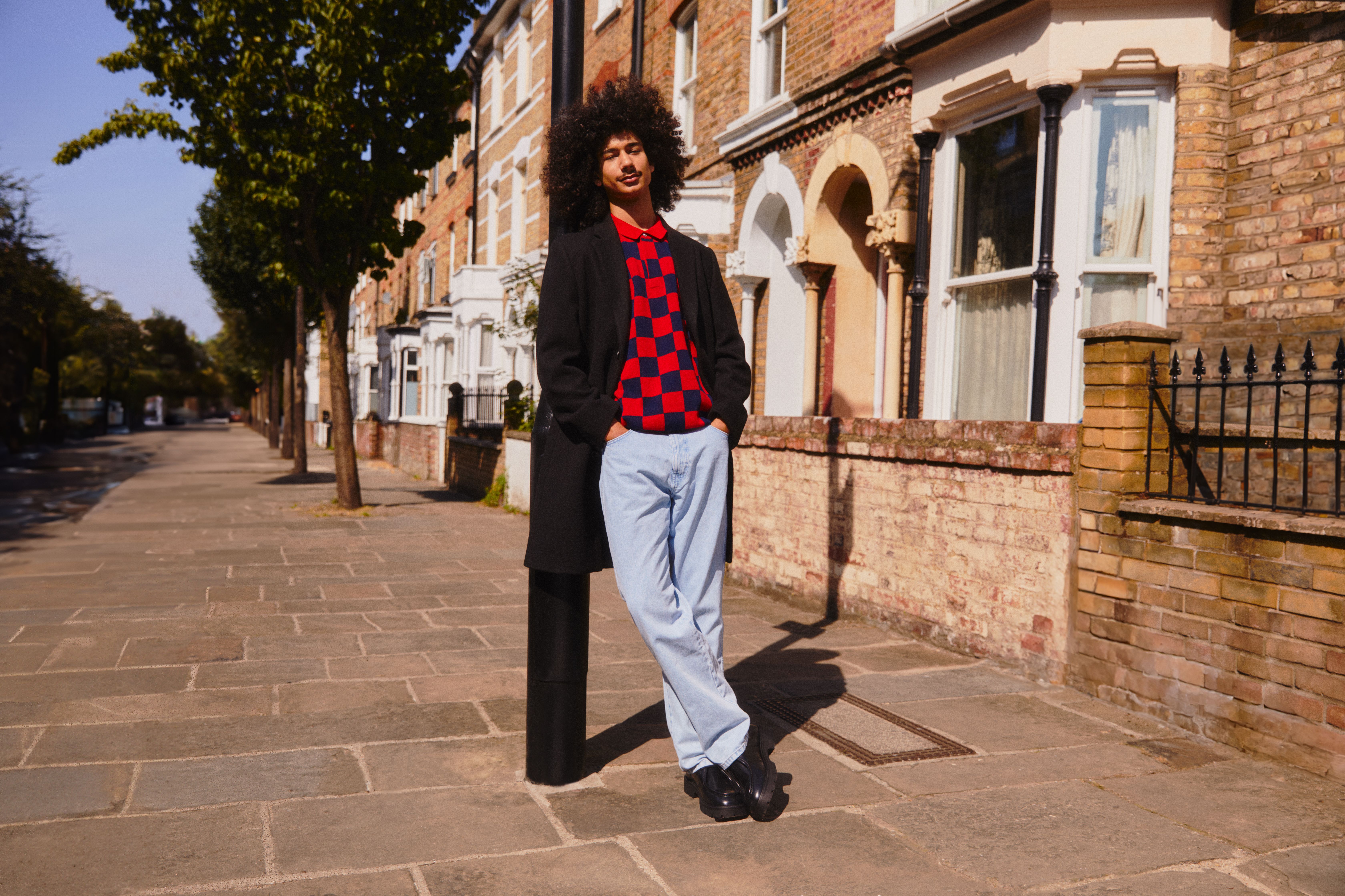 Mann, der sich an eine Straßenlaterne lehnt, trägt einen leuchtenden Pullover und helle Jeans, die sich vom Hintergrund abheben